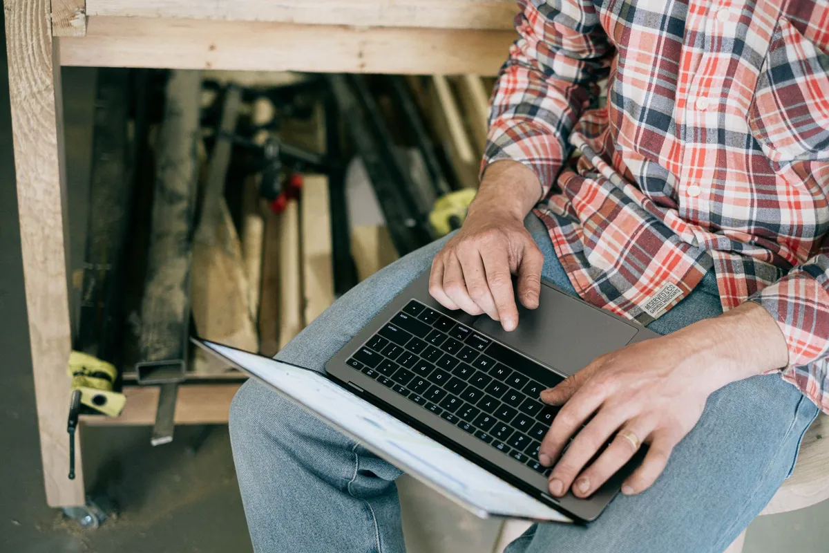 A craftsperson in a workshop working on a laptop surrounded by tools